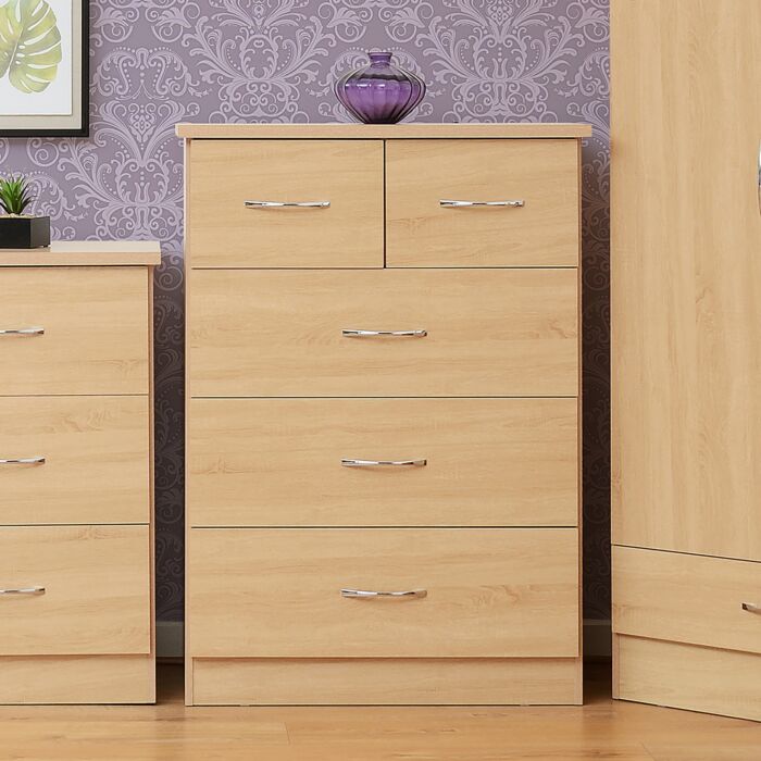 100-102-114_1 - Jamies Furniture Tall light-wood dresser with five drawers and metal handles on a wooden floor, purple patterned wallpaper behind; a purple glass vase sits on top.