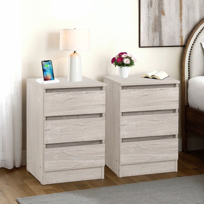 Two light wood nightstands with three drawers each in a bedroom; left one has a lamp and smartphone, right one has flowers and an open book.